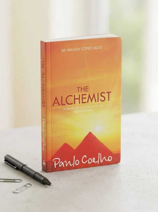 Book titled 'The Alchemist' by Paulo Coelho on a white surface with a pen and paper clips.
